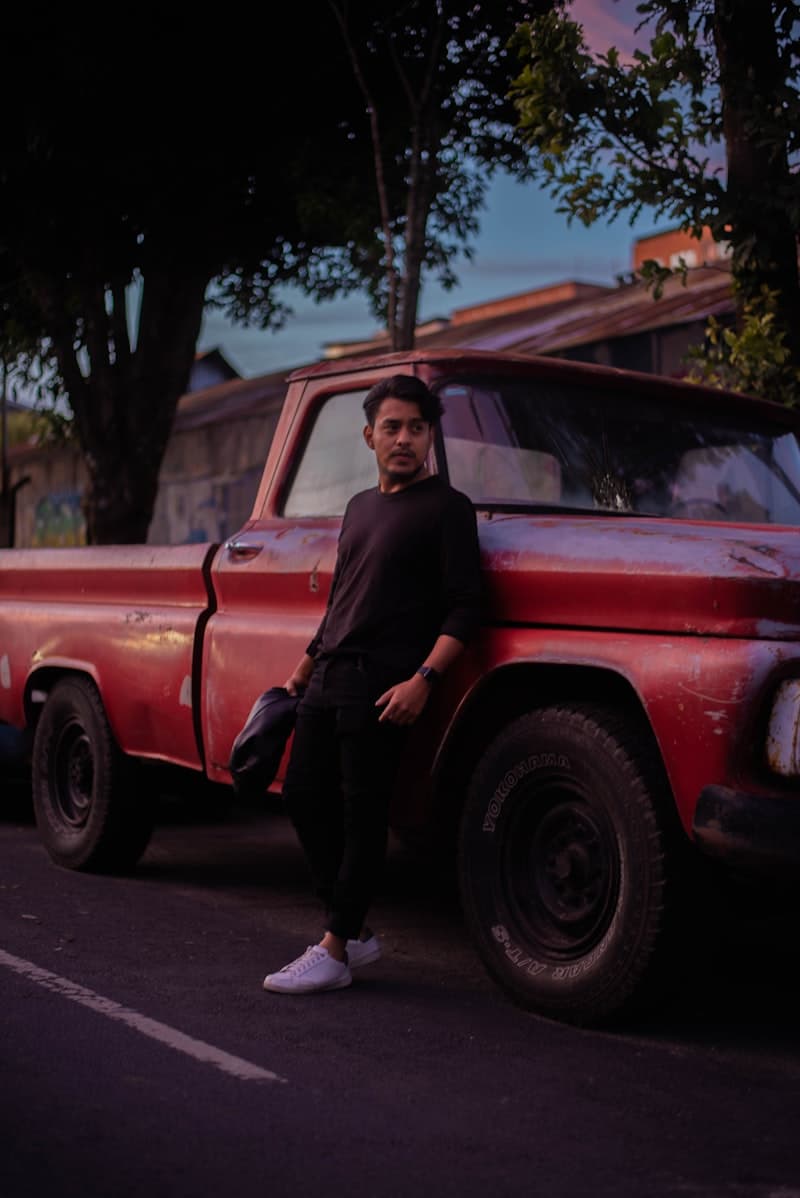 A man leaning against a red pickup truck at dusk