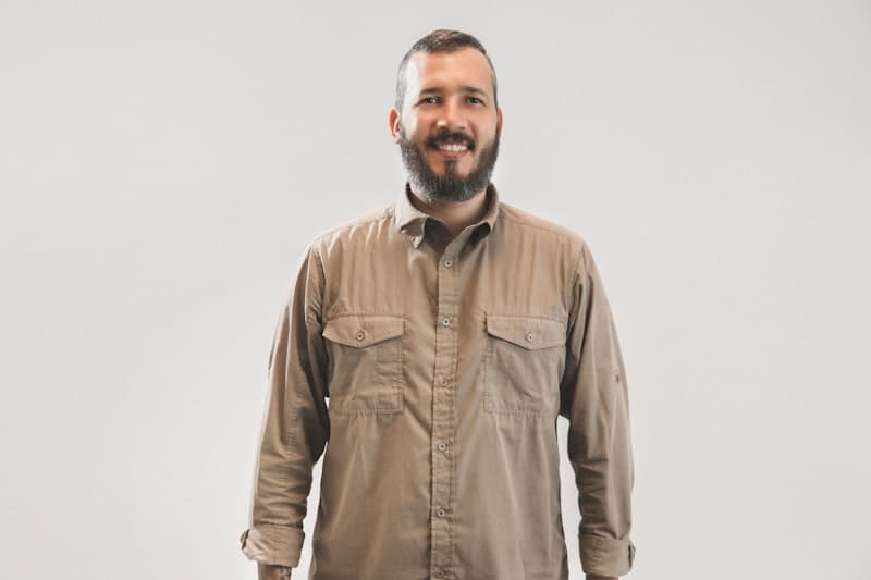 A bearded man in a khaki work shirt against a plain wall