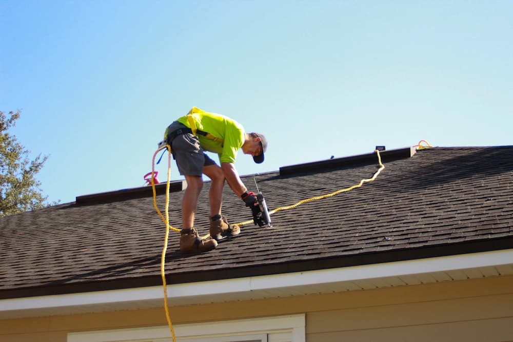 A roofer fastening shingles with a power drill