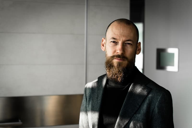 A bearded man in a dark jacket standing in a plain room