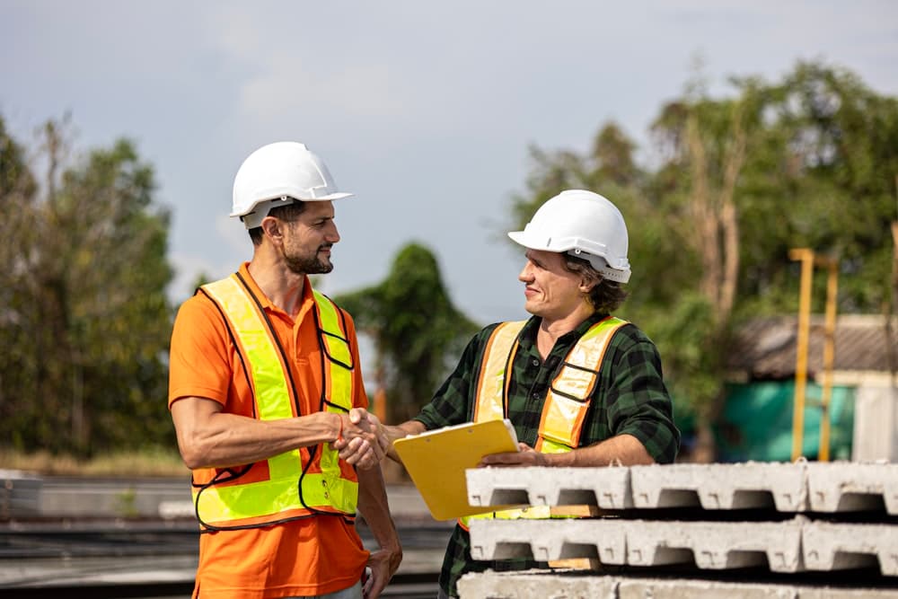 Two workers in safety vests shaking hands over stacked materials